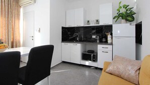 Cozinha compacta com armários brancos, bancada preta e micro-ondas. Há uma área de refeições com mesa e cadeiras, e um sofá com almofada.