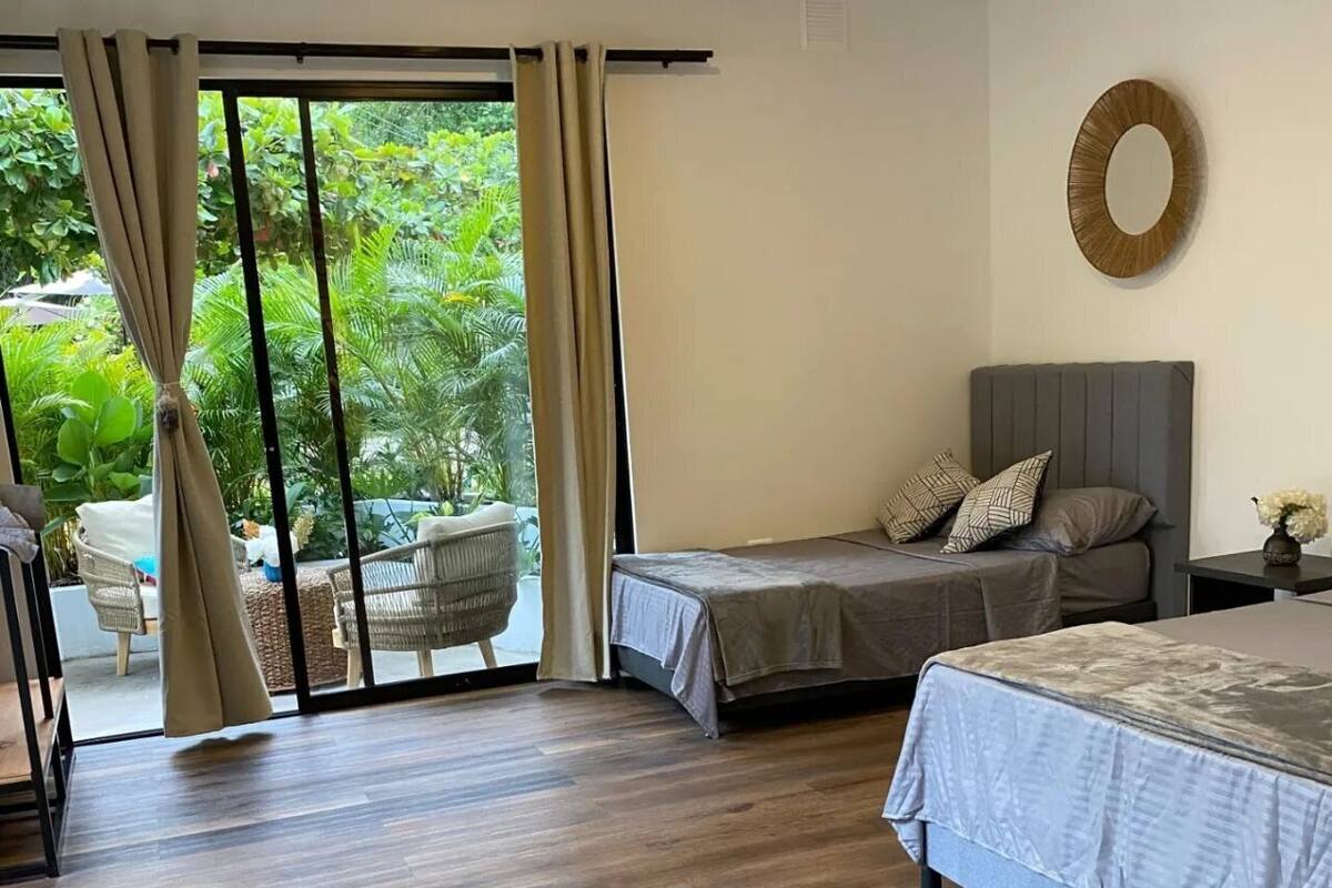 Um quarto com uma grande porta de correr de vidro que dá para um pátio, uma cama com cabeceira cinza e uma pequena mesa com um vaso de flores.
