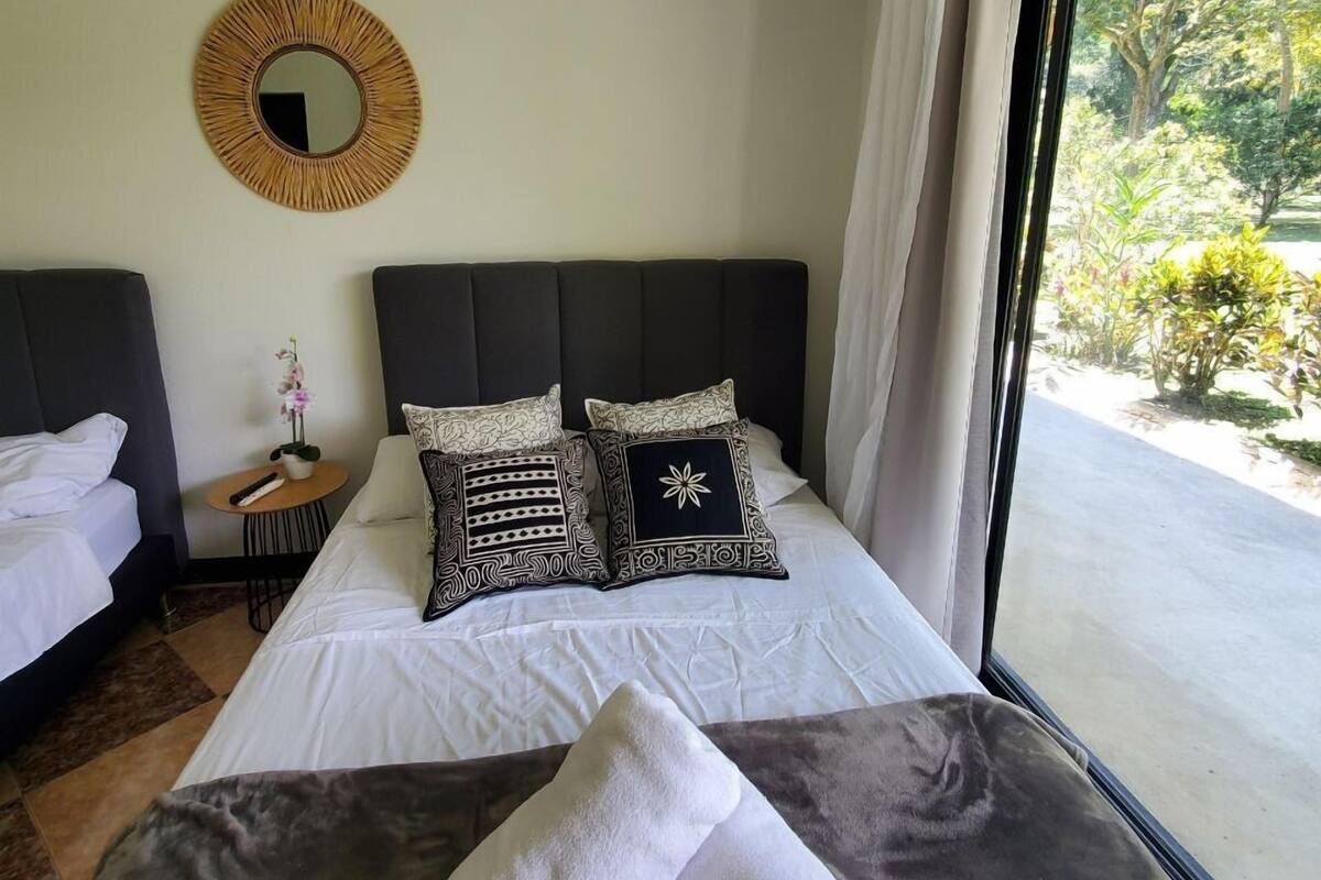 Quarto de hotel com uma cama grande, duas mesas de cabeceira, um espelho redondo e vista para o verde lá fora.
