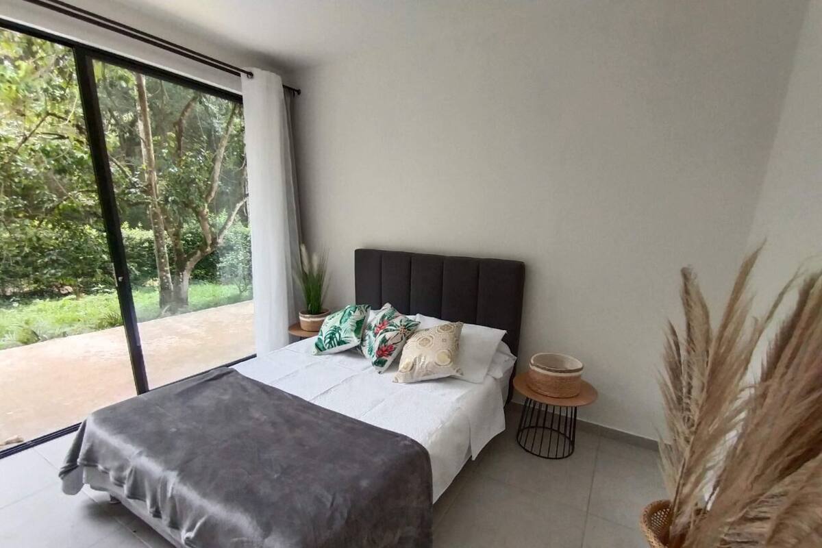 Quarto moderno com uma cama grande, vista para o verde através de uma porta deslizante e uma pequena mesa de cabeceira com uma tigela e uma planta.