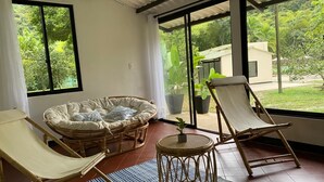 Een zonnige kamer met een groot raam, een rieten bank, twee fauteuils en een klein rond tafeltje.