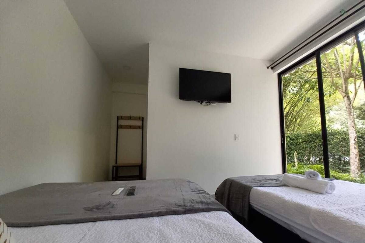 Quarto com duas camas, televisão de tela plana fixada na parede e janela com vista para área verde.