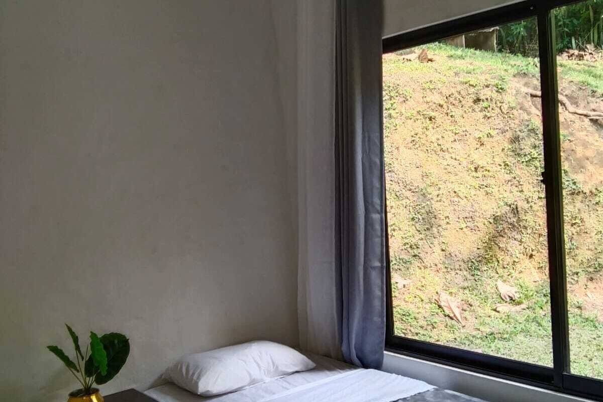 Um quarto pequeno e simples com uma cama de solteiro, uma mesa de cabeceira com uma planta e uma janela com vista para uma colina.