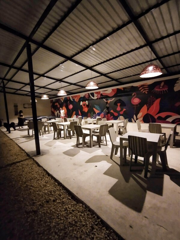 An indoor dining area with tables and chairs, a mural on the wall, and a metal roof structure.