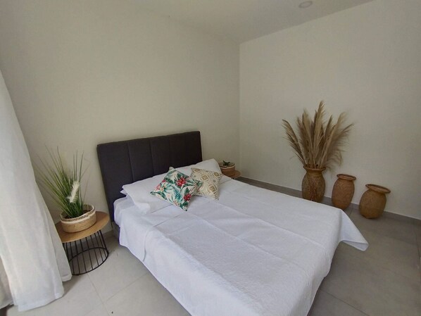 A neatly made bed with white linens, a dark headboard, and a floral pillow. There are three potted plants on the floor beside the bed.