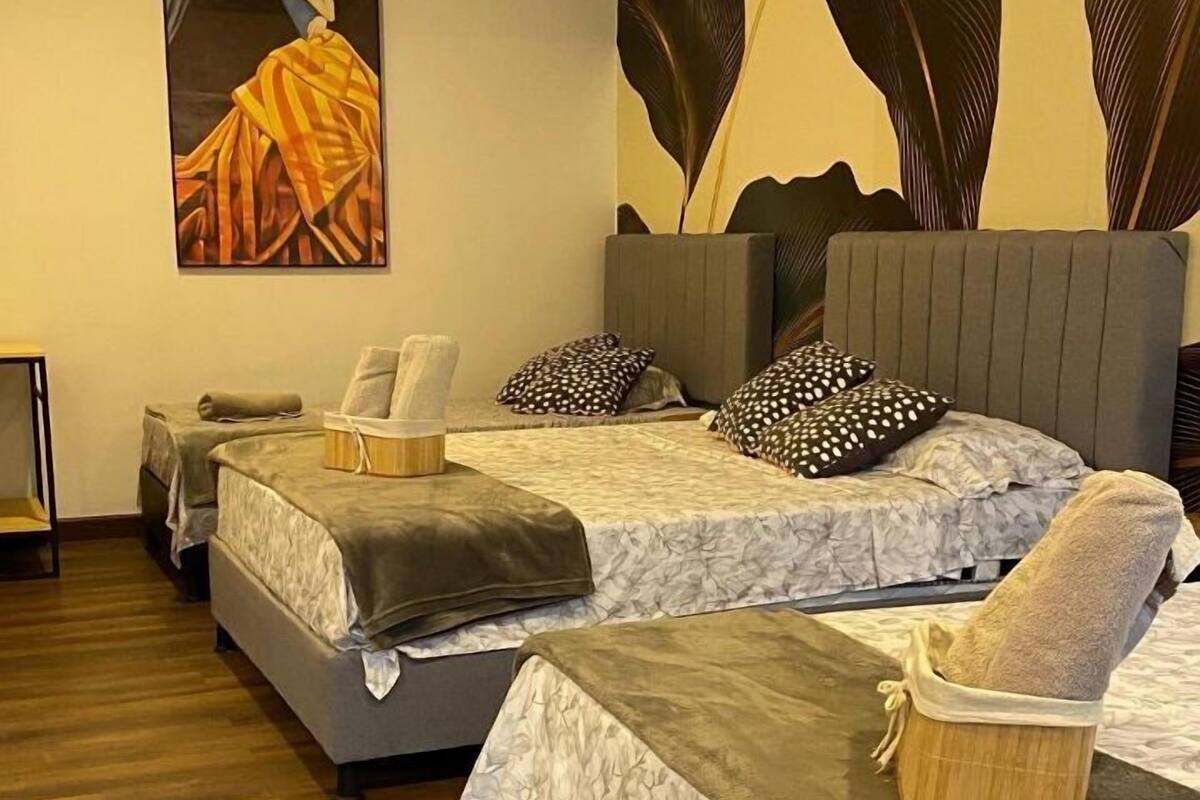 Quarto de hotel com três camas, cada uma com cabeceira cinza e roupa de cama com padrão floral. Um quadro abstrato grande está na parede acima das camas.