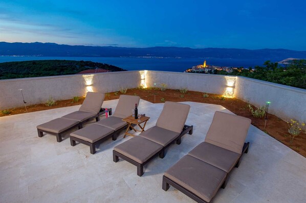 A rooftop terrace with six lounge chairs and a small table, overlooking a coastal cityscape at dusk.