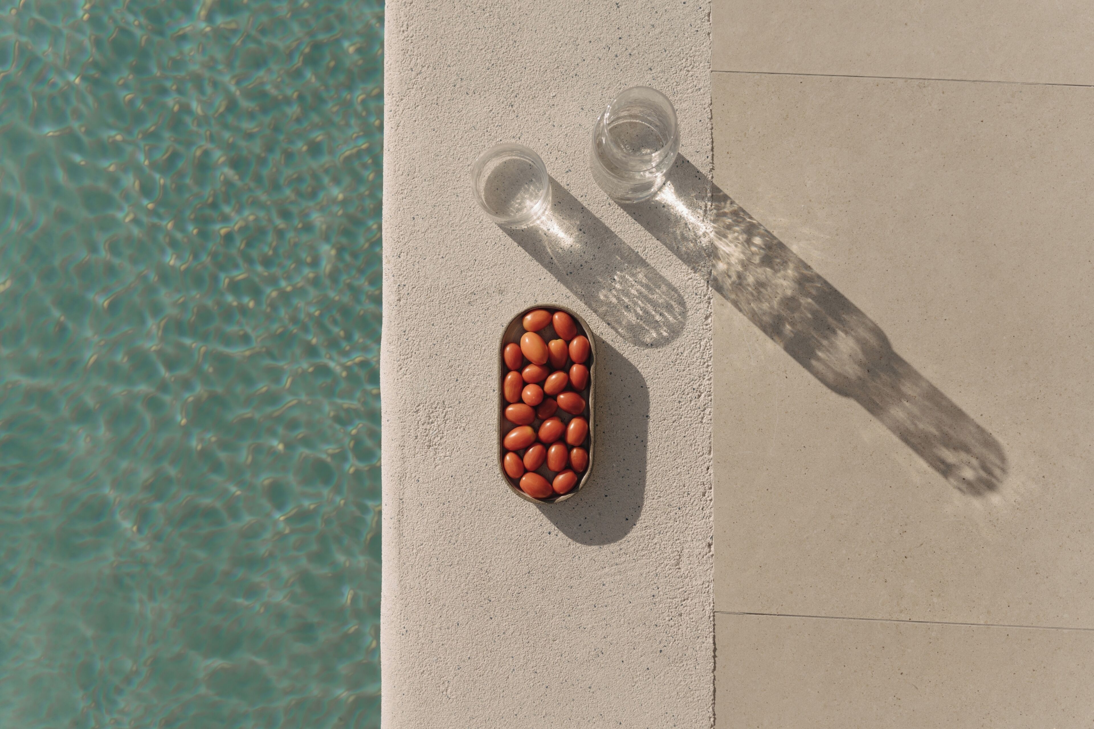 A poolside area with a concrete edge, two empty glasses, and a bowl of orange objects.