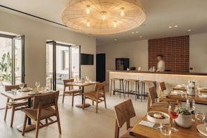 A modern dining area with wooden furniture, a central light fixture, and a kitchen counter in the background. - Bard De Sol (Mykonos)