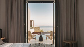 A bedroom with a bed, a small table, two chairs, and a view of the sea through an open door. - Bard De Sol (Mykonos)