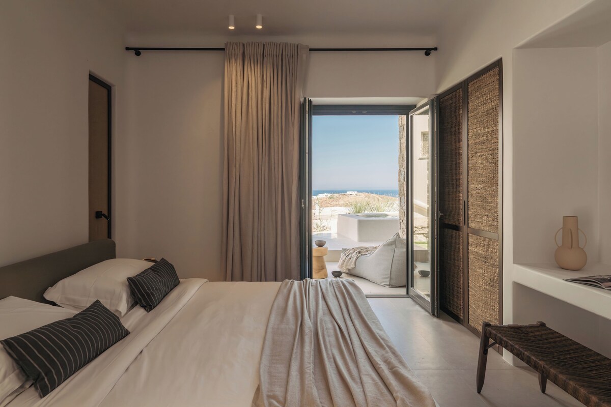 A modern bedroom with a large bed, a view of the outdoors, and a wooden bench.