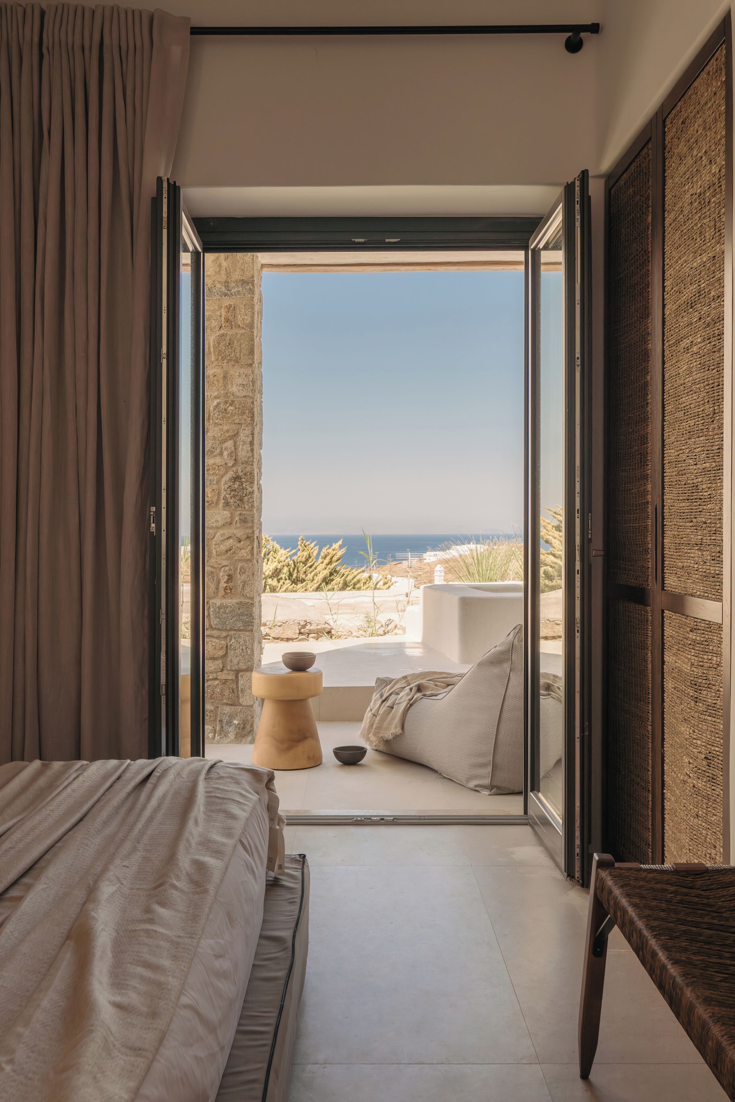 A bedroom with a bed, a bench, and a view of the beach and ocean through an open door.