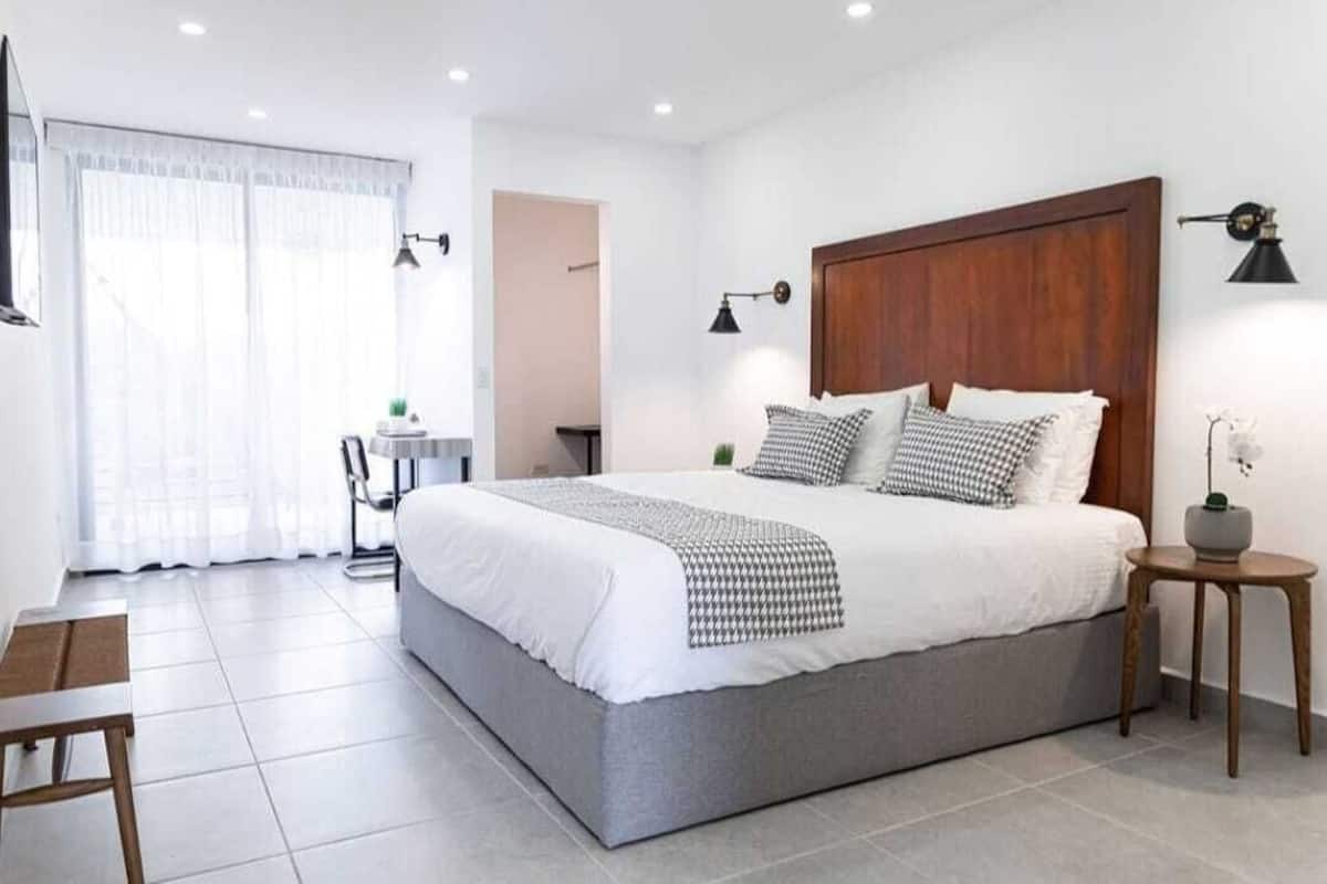 Ein modernes Schlafzimmer mit einem großen Bett, einem kleinen Tisch und einem Sessel. Ein Fenster mit Vorhängen und eine Wandlampe sind vorhanden.
