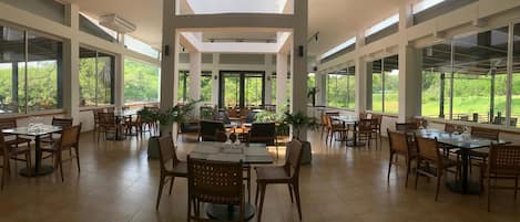 A spacious dining area with high ceilings, large windows, and natural light. Tables are set with chairs and centerpieces.