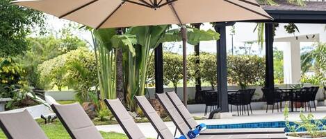 Una hilera de sillas de playa con cojines color beige y toallas azules, protegidas por grandes sombrillas color beige, en una zona junto a la piscina con una pileta y plantas tropicales al fondo.