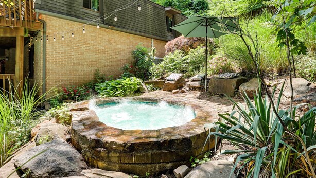 A backyard with a hot tub surrounded by rocks and plants, a wooden deck, and a brick house. - Spacious King of Prussia Home w/ 2 Game Rooms (King of Prussia)