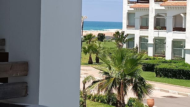 On peut admirer un bâtiment blanc doté d’un balcon qui donne sur une plage, ainsi qu’un jardin bien entretenu avec des palmiers.