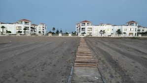 Une propriété située en bord de mer, composée de plusieurs bâtiments blancs, d’une plage de sable et d’une promenade en bois menant aux bâtiments.