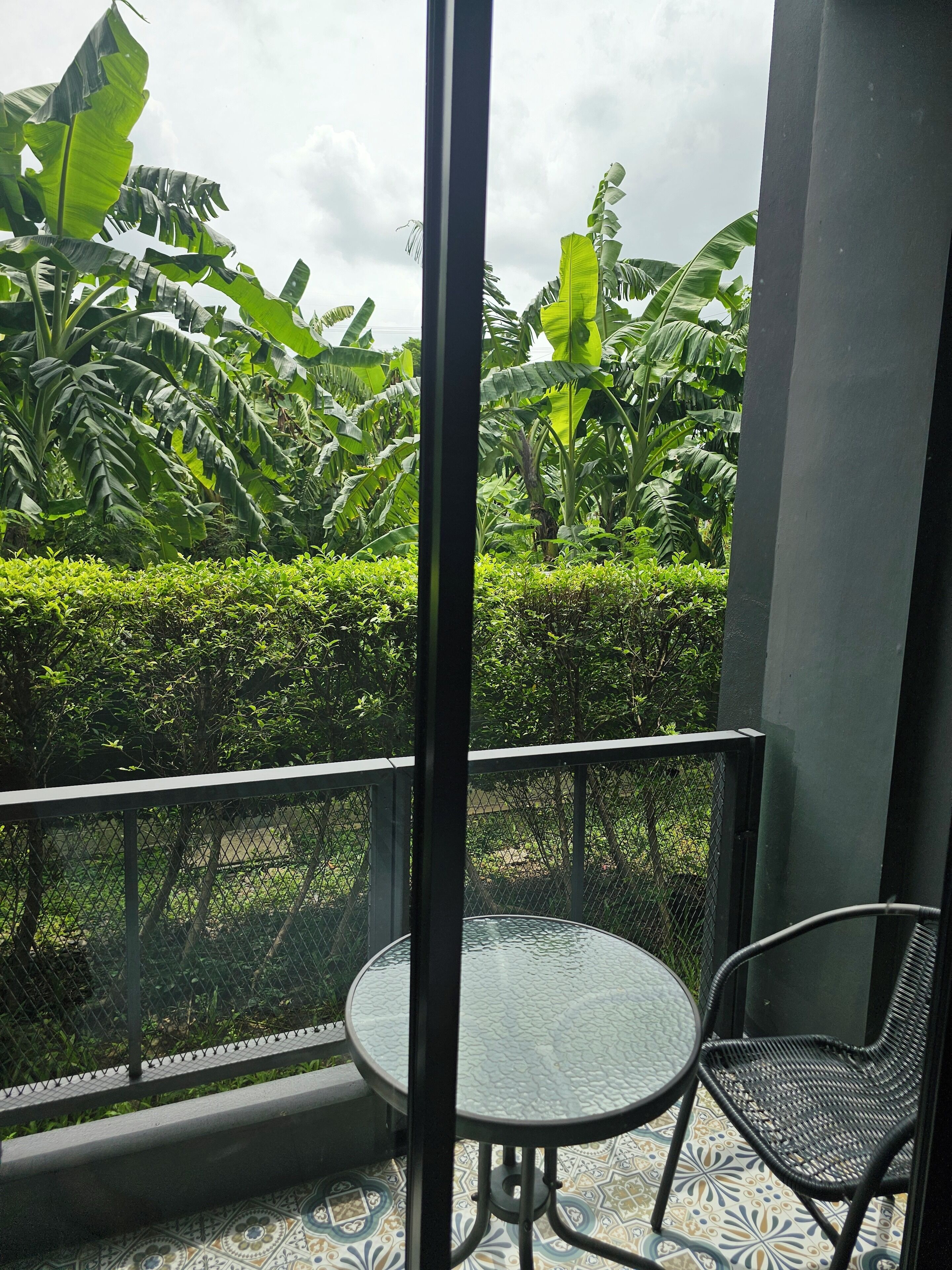A balcony with a glass table and chair, surrounded by lush greenery and a patterned floor.
