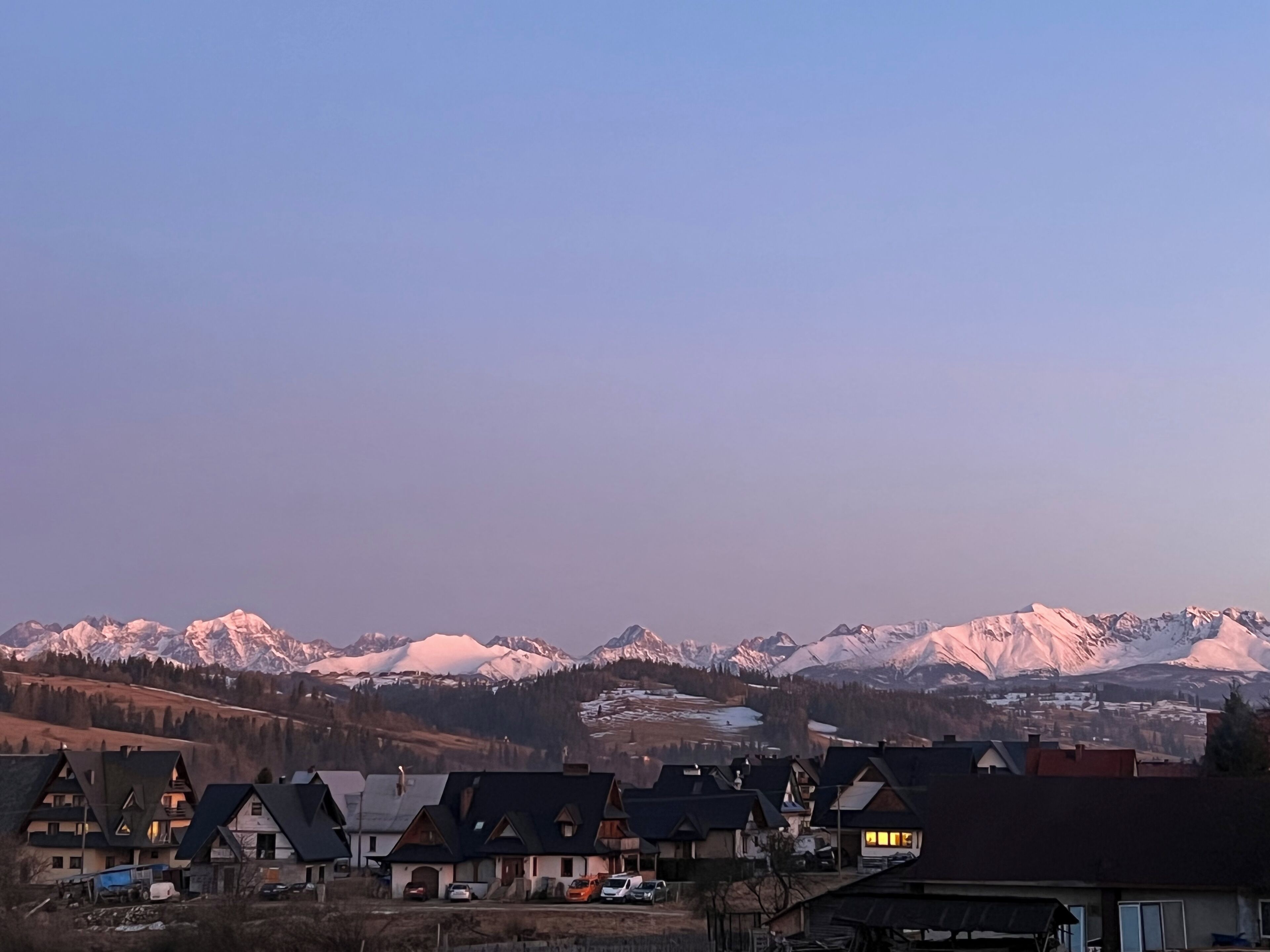 A mountain village with traditional chalet-style houses, a clear sky, and snow-capped mountains in the background.