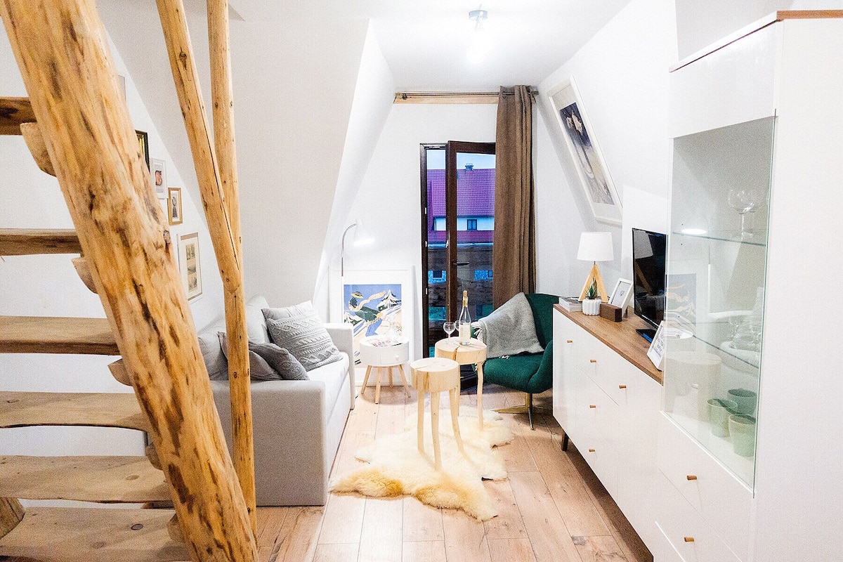 Una sala moderna con un sofá gris, un sillón verde, una mesa de centro de vidrio y un manto blanco de piel. A la izquierda, una escalera de madera; a la derecha, un aparador de vidrio. Se ve un edificio a través de una ventana.