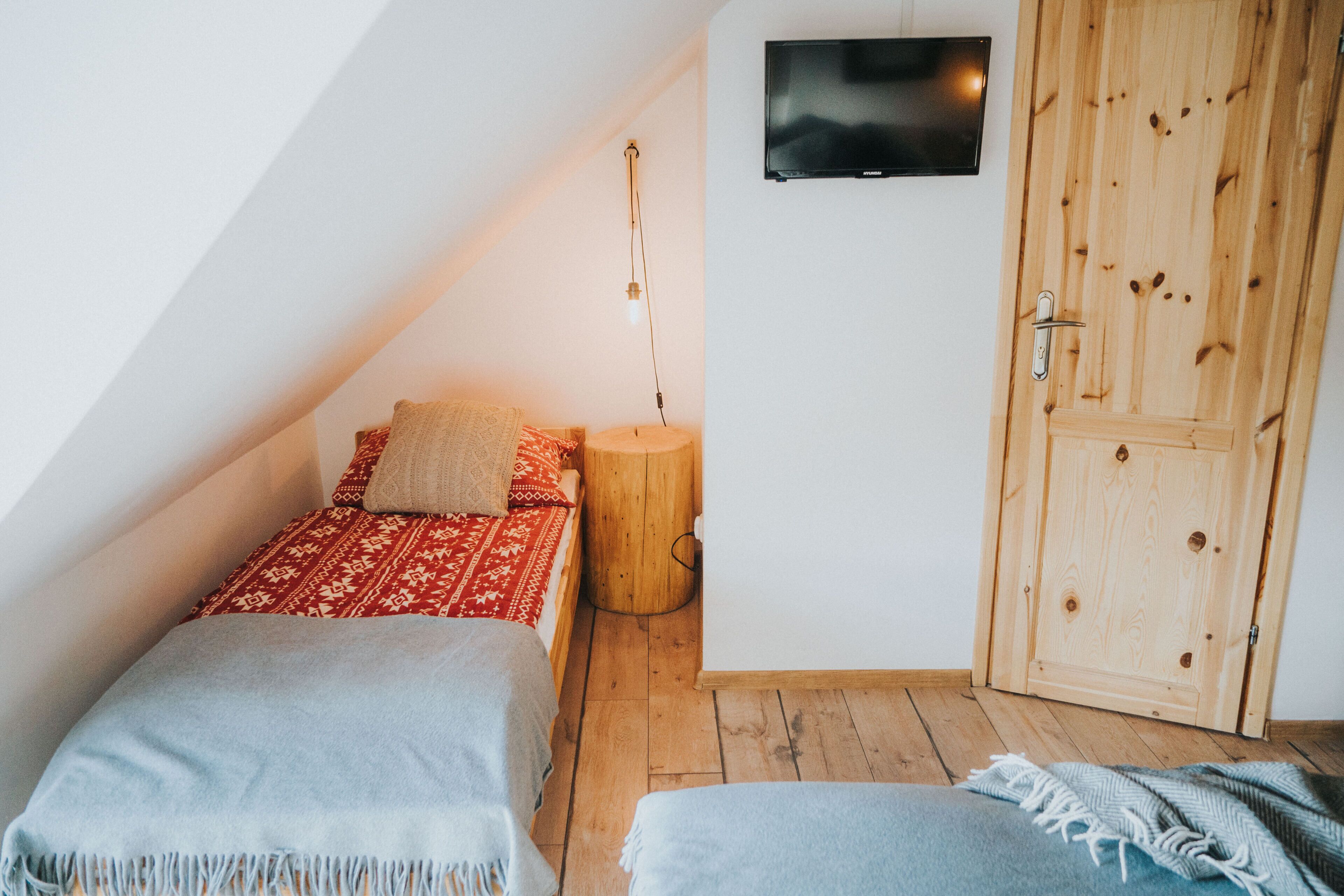 Una habitación pequeña y acogedora, con techo inclinado, un armario de madera, una cama con ropa de cama estampada, una mesita de noche con lámpara, y un televisor montado en la pared.