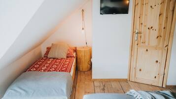 Una habitación pequeña y acogedora, con techo inclinado, un armario de madera, una cama con ropa de cama estampada, una mesita de noche con lámpara, y un televisor montado en la pared.