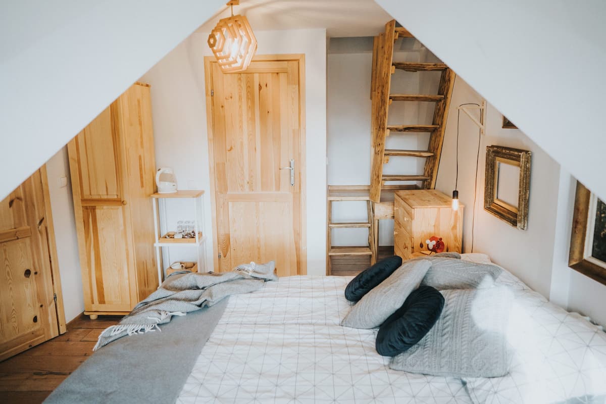 Un dormitorio con techo inclinado, armario de madera, cama con almohadas, mesita de noche y una escalera.