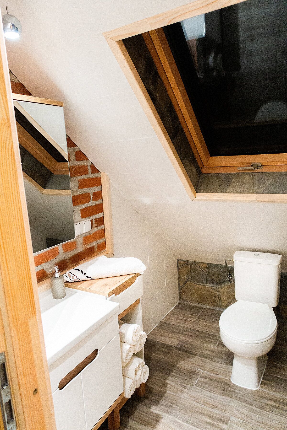 A bathroom with a skylight, white fixtures, a brick wall, and a wooden vanity.