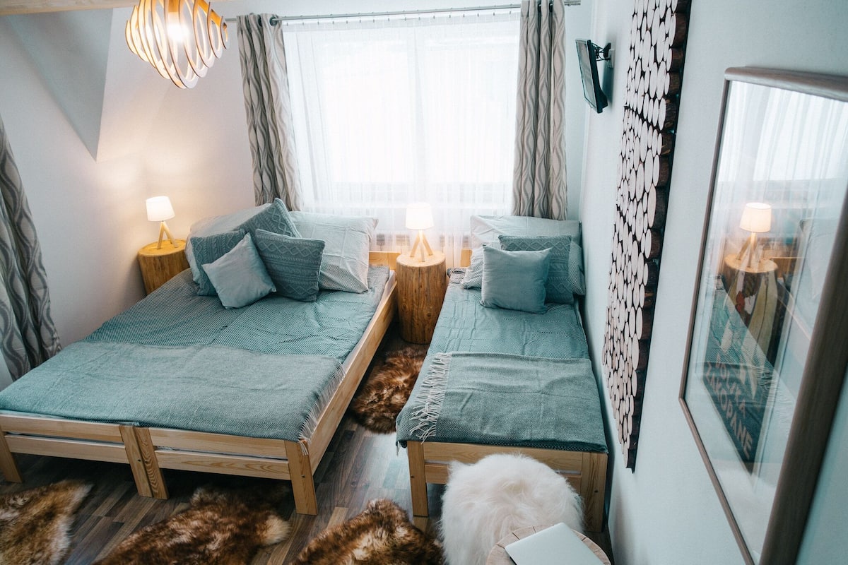 Una habitación con dos camas de madera, una mesita de noche de madera con una lámpara, una alfombra de piel y un adorno en la pared.