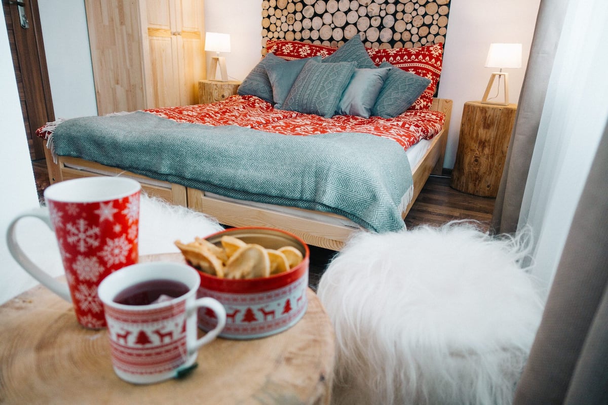 Un dormitorio acogedor con una cama de madera, una colcha estampada en rojo y blanco, un almohadón blanco mullido y una mesita de noche de madera con una lámpara.