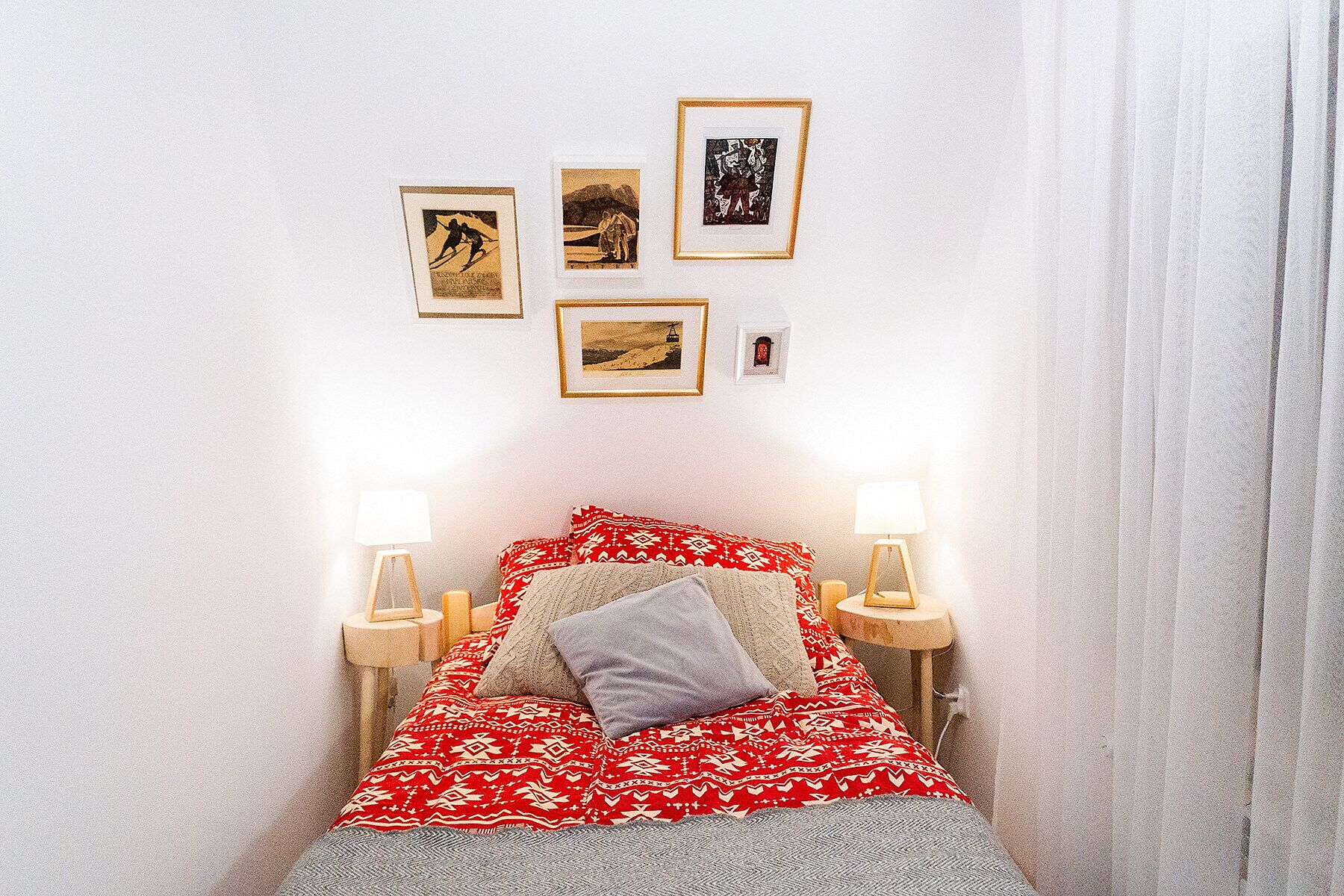 Uma cama com um cobertor vermelho estampado, um travesseiro cinza e duas luminárias de cabeceira. Quatro quadros emoldurados estão pendurados na parede acima da cama.