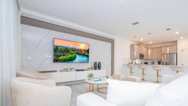 Ein modernes Wohnzimmer mit Flachbildfernseher, weißen Sofas, einem runden Couchtisch und einer Küchenzeile mit weißen Schränken und Edelstahlgeräten.