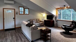 Un soggiorno in stile loft con soffitto alto, un divano, una poltrona, un televisore e una finestra vista alberi.