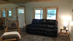 A cozy living room with a black leather sofa, a wooden rocking chair, and a small rug. There are two windows with wooden frames, a door, and a small table with a lamp. - Whitetail Hollow  (McBain)