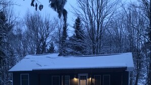 A single-story cabin with a snow-covered roof, a front porch, and a door with windows on either side. - Whitetail Hollow  (McBain)