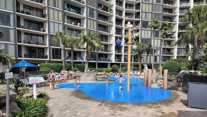 Área de parque aquático com piscina, espreguiçadeiras e um edifício alto ao fundo.