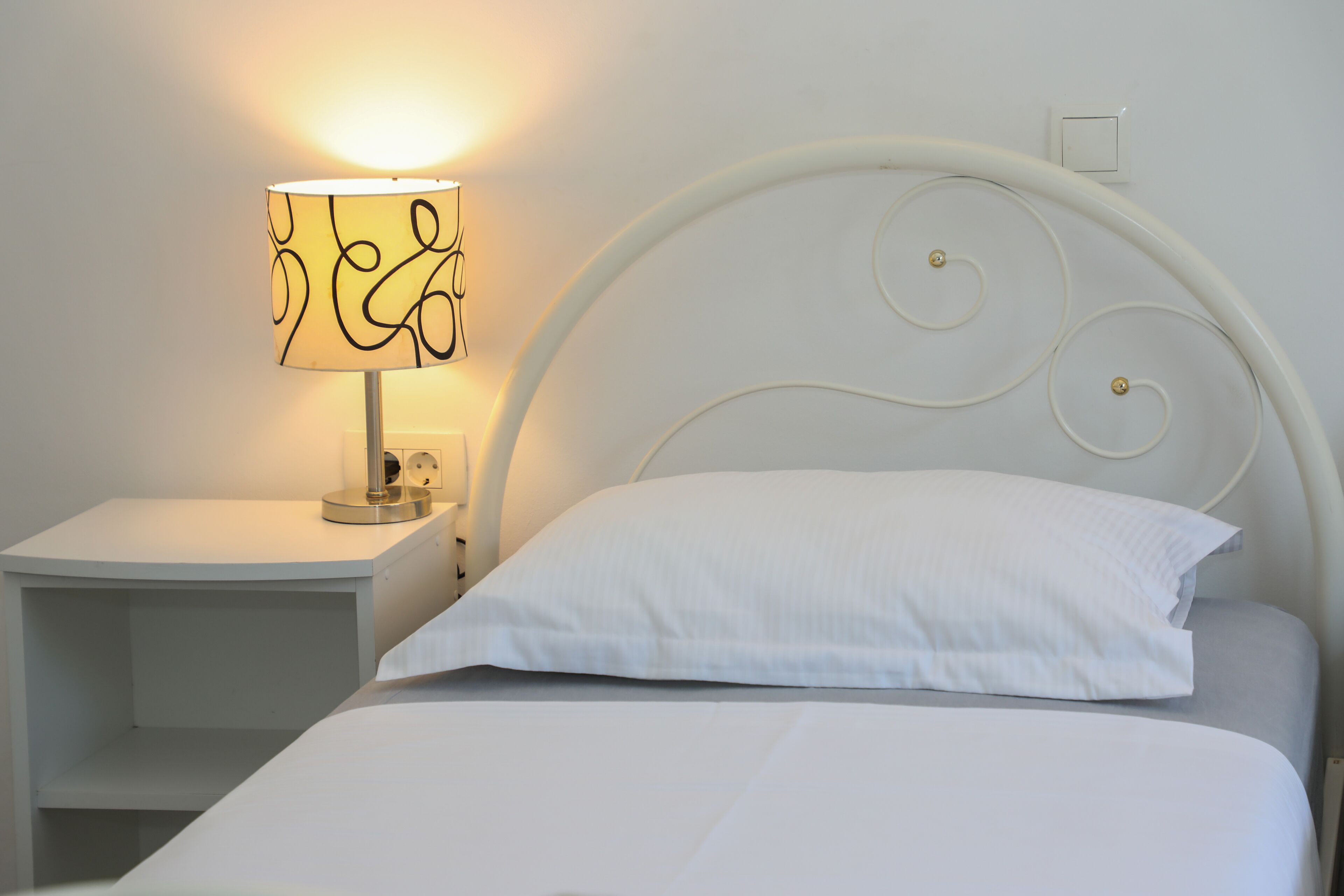 A neatly made bed with a white pillow and a white headboard. There is a bedside table with a lamp and a power outlet.