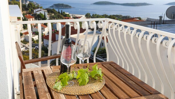 Et træbord med en vævet bordskåner, to vinglas og en vinflaske på en balkon med udsigt over kysten.