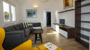 En moderne stue med en sofa, en gul lænestol, et lille bord med glas og en bog, et fjernsyn og et maleri på væggen.