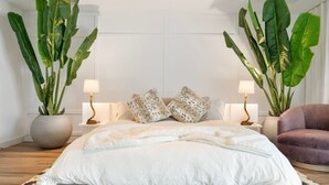 Quarto com cama grande, duas mesas de cabeceira com luminárias, uma cadeira e duas plantas em vasos grandes.