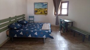 Un dormitorio con cama, escritorio, silla, banco, un cuadro y una ventana.