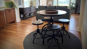 Ampla sala de estar com uma mesa redonda de madeira, dois banquinhos, um sofá e um ventilador de teto.