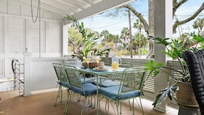 A covered patio with a table and chairs, surrounded by potted plants and a view of palm trees. - Hemingway | Jungle Hideaway (Folly Beach)