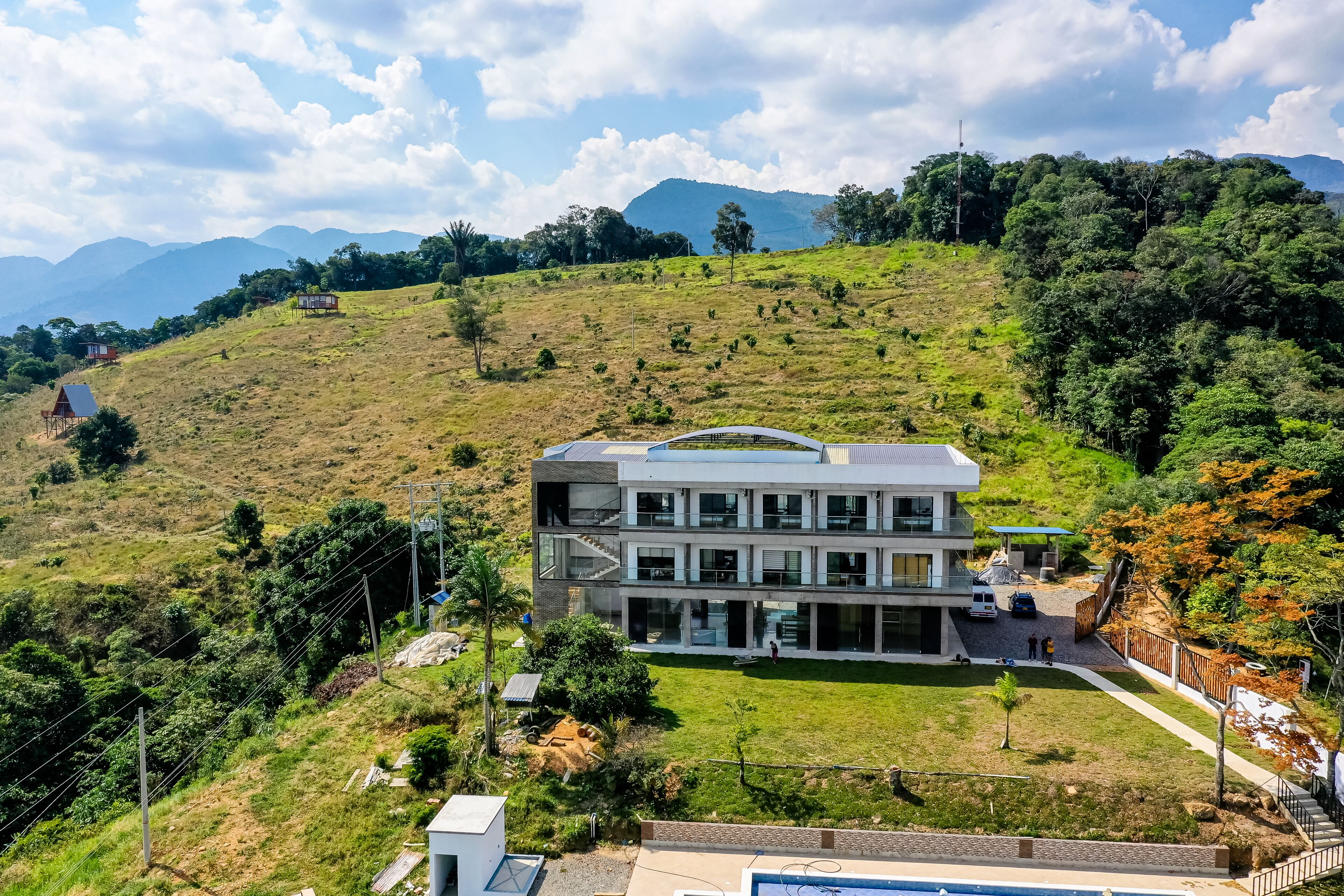 A modern multi-story building with a flat roof and large windows, surrounded by a lush green landscape and hills in the background.