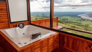 A wooden bathroom with a large bathtub and a view of a green landscape through a large window.