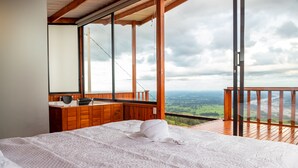 A bedroom with a large bed, a wooden dresser, and a view of the landscape through a balcony.