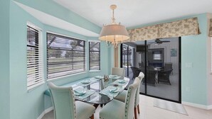 Un comedor con una mesa de vidrio, cuatro sillas y una lámpara de araña. Hay dos ventanas con persianas y una puerta corrediza de vidrio que da a una cocina.