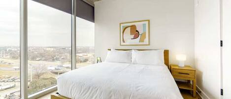 A modern bedroom with a large bed, a nightstand, a lamp, and a city view through large windows.