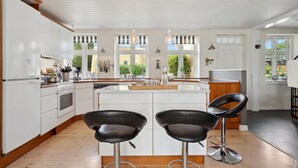 A modern kitchen with white cabinetry, a central island, two black bar stools, and a view of greenery outside. - 4 bedroom nice home in Nyborg (Nyborg)